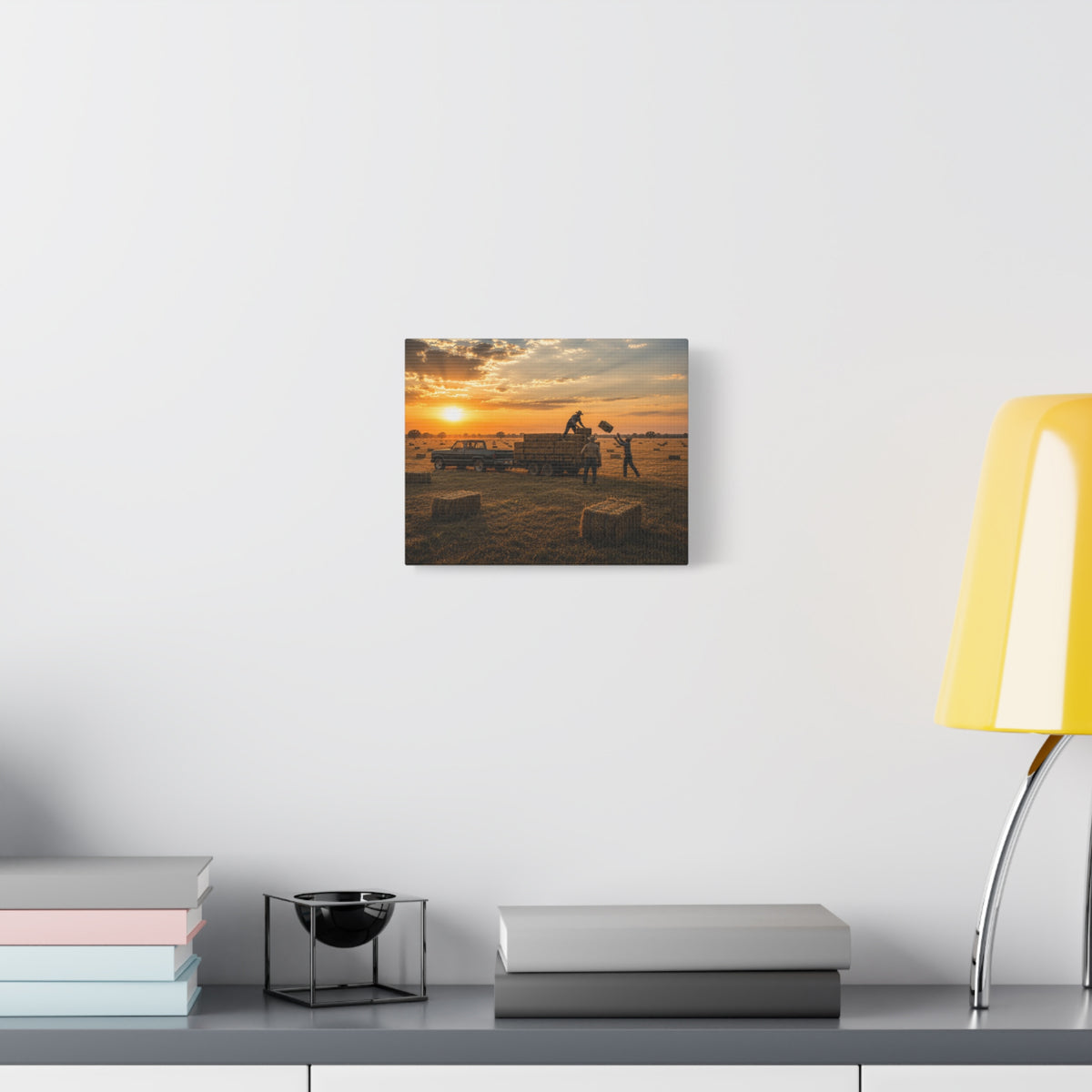 A square Hay Season canvas print showing farmworkers loading rectangular hay bales onto a flatbed trailer beside a pickup truck in a golden field at sunset, displayed on a white wall above books and a yellow lamp.