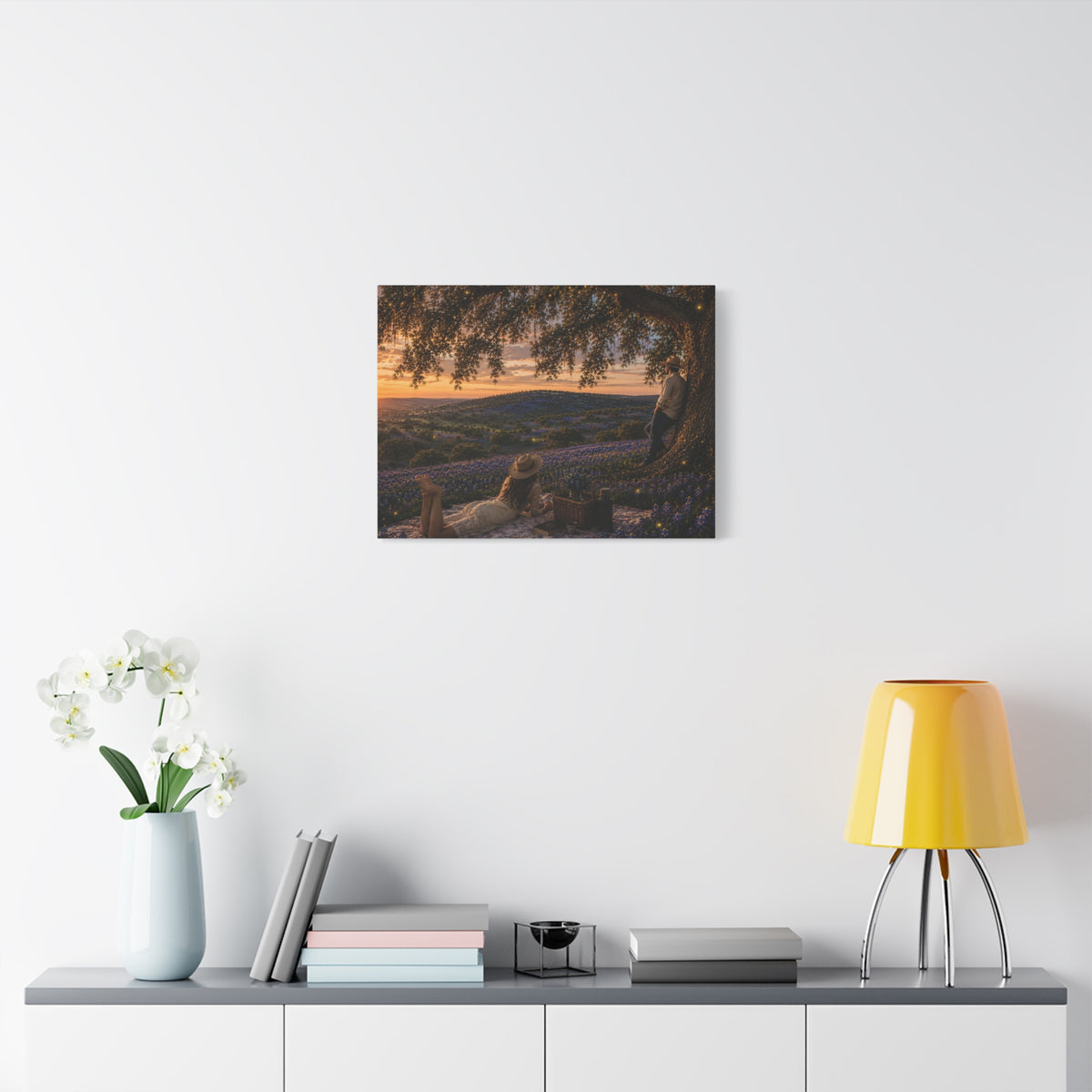 A framed wall art print depicting a sunset landscape with purple lavender fields and a tree, displayed on a white wall above a console table with a vase of white flowers, books, a small decor object, and a yellow lamp.