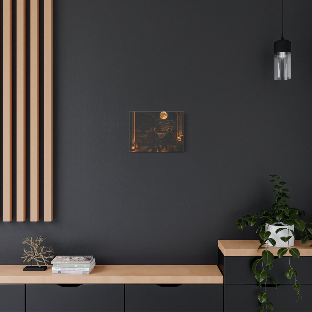 A Harvest Moon canvas print showing an older couple holding hands in rocking chairs on a farmhouse porch with a sleeping dog, glowing lanterns, and a full moon over the fields, displayed on a dark gray wall above a wooden console with a plant and hanging light.
