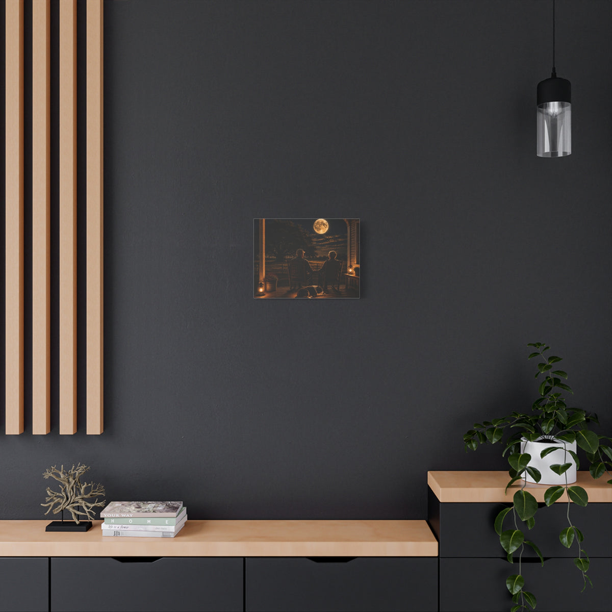 A Harvest Moon canvas print showing an older couple holding hands in rocking chairs on a farmhouse porch with a sleeping dog, glowing lanterns, and a full moon over the fields, displayed on a dark gray wall above a wooden console with a plant and hanging light.