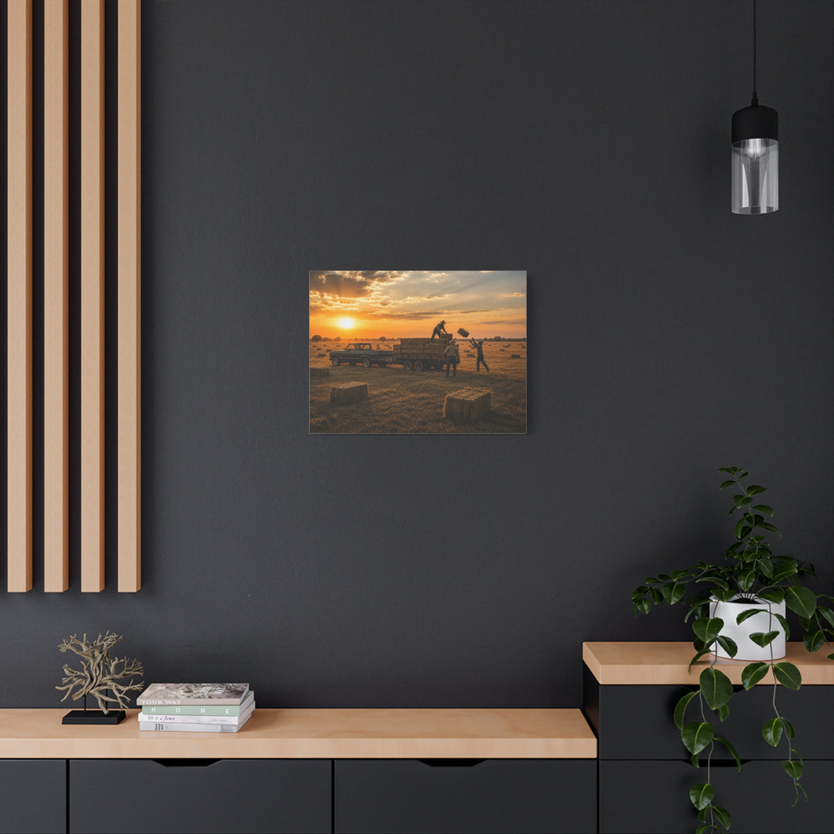 A square Hay Season canvas print showing farmworkers loading rectangular hay bales onto a flatbed trailer beside a pickup truck in a golden field at sunset, displayed on a dark charcoal wall above a light wood console with books and a plant.