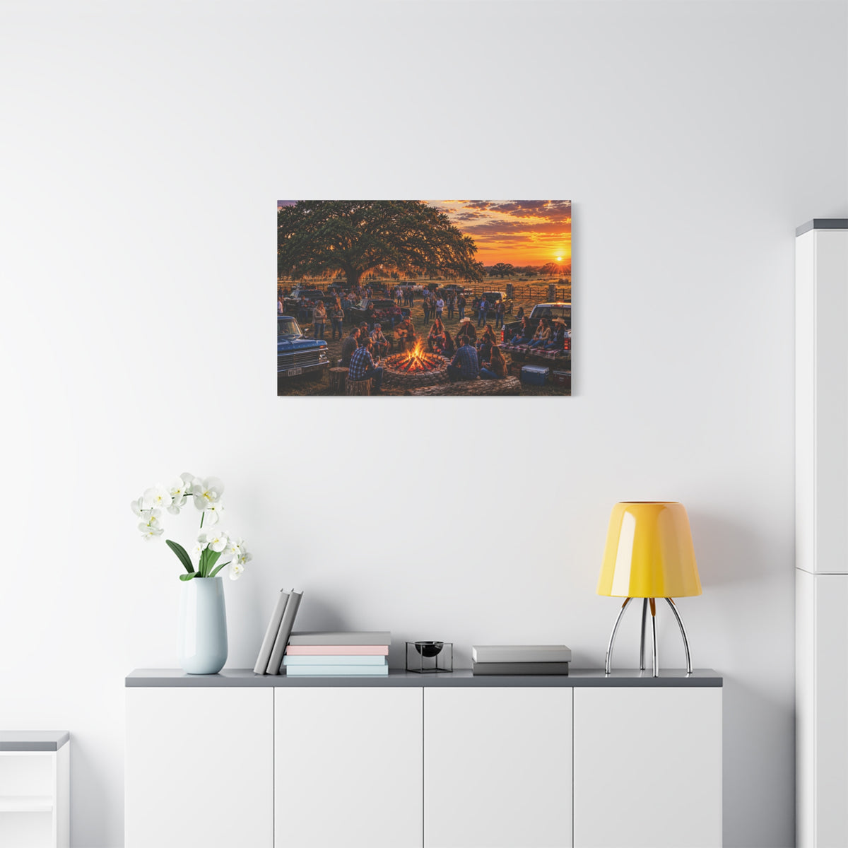 A rectangular Campfire Stories canvas print showing a large group gathered around a ranch campfire beneath an oak tree at sunset, displayed above a white cabinet with a yellow lamp and white flowers.