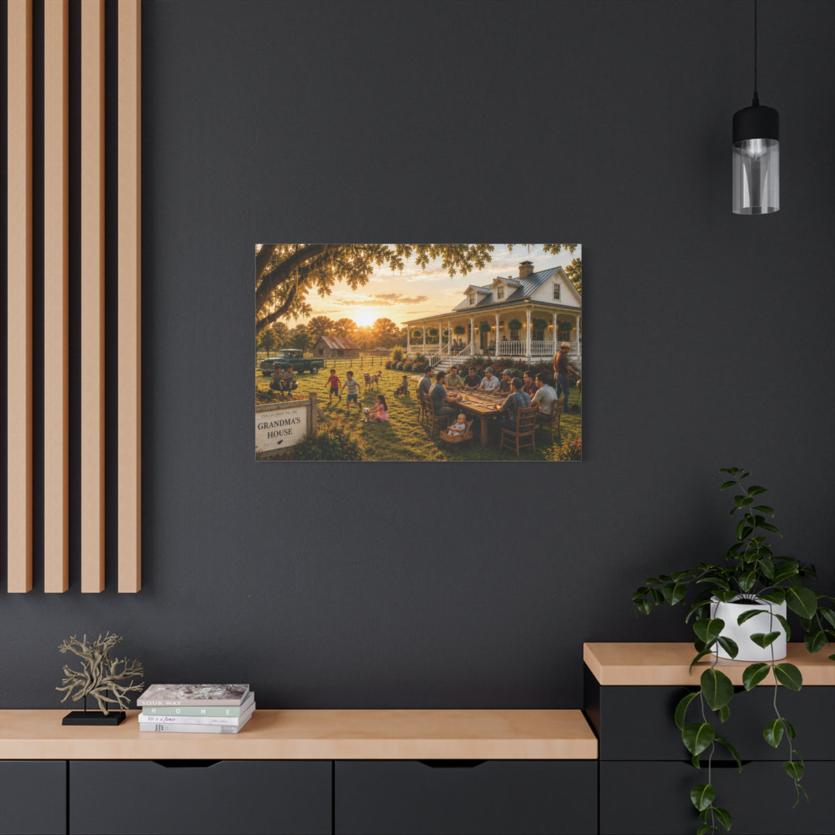 A Grandma's House canvas print showing a warm family gathering outside a white farmhouse at sunset, displayed above a white cabinet with a yellow lamp and white flowers.