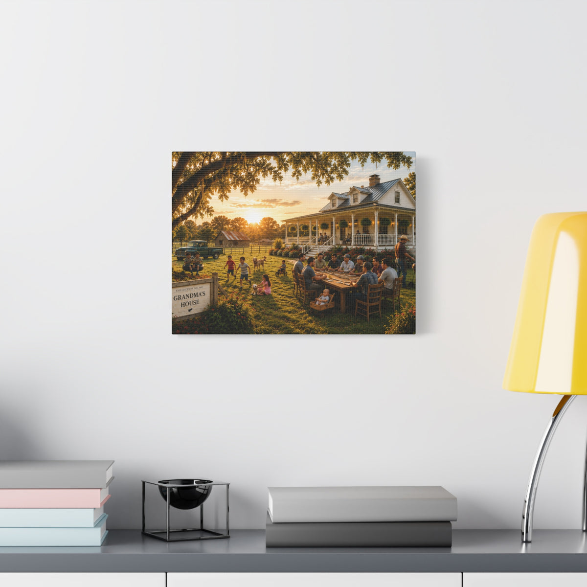 A rectangular Grandma's House canvas print showing a family gathering outside a farmhouse at sunset, displayed on a light gray wall above books and a yellow lamp.