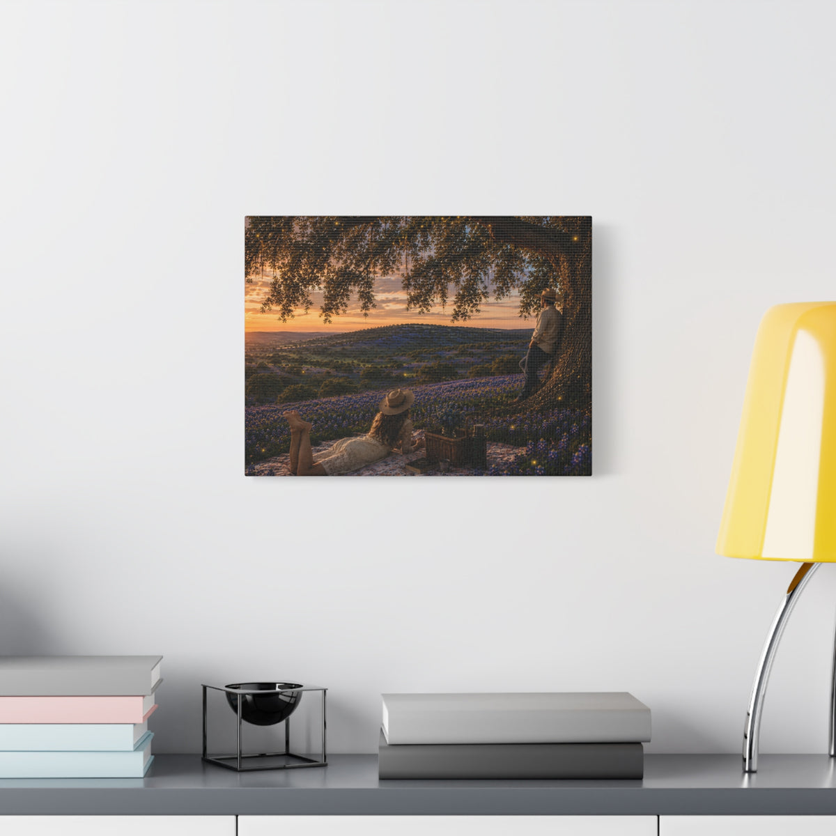 A framed wall art print depicting a scenic landscape with an olive tree, a reclining figure, and a sunset over purple flowers, displayed on a white wall above a shelf with books and a yellow lamp.