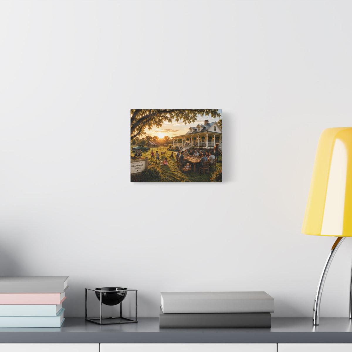 A square Grandma's House canvas print showing a warm family gathering outside a farmhouse at sunset, displayed on a white wall above books and a yellow lamp.