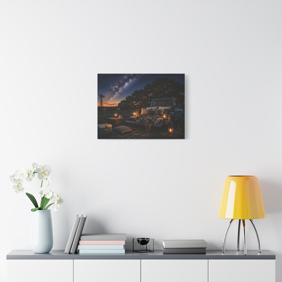 A Starry Night canvas print showing a cozy outdoor bed beside a vintage pickup in a country field, with lanterns, pillows, blankets, windmills, and the Milky Way overhead, displayed on a white wall above a console with a yellow lamp, books, and white flowers.