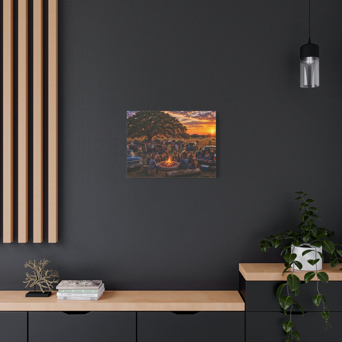 A square canvas art print depicting a lively campfire gathering under a warm sunset sky, displayed on a dark charcoal wall above a light wood console table with a potted plant and books.