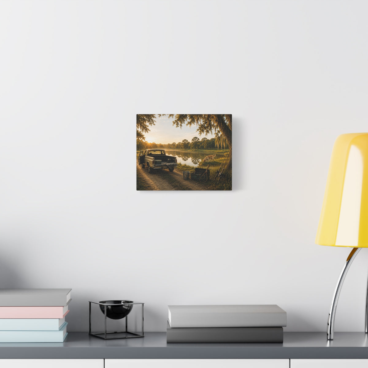 A square The Old Fishing Hole canvas print showing a vintage pickup parked beside a quiet pond at sunset, with a dog, fishing rods, tackle, a cooler, and a moss-draped tree, displayed on a white wall above books and a yellow lamp.