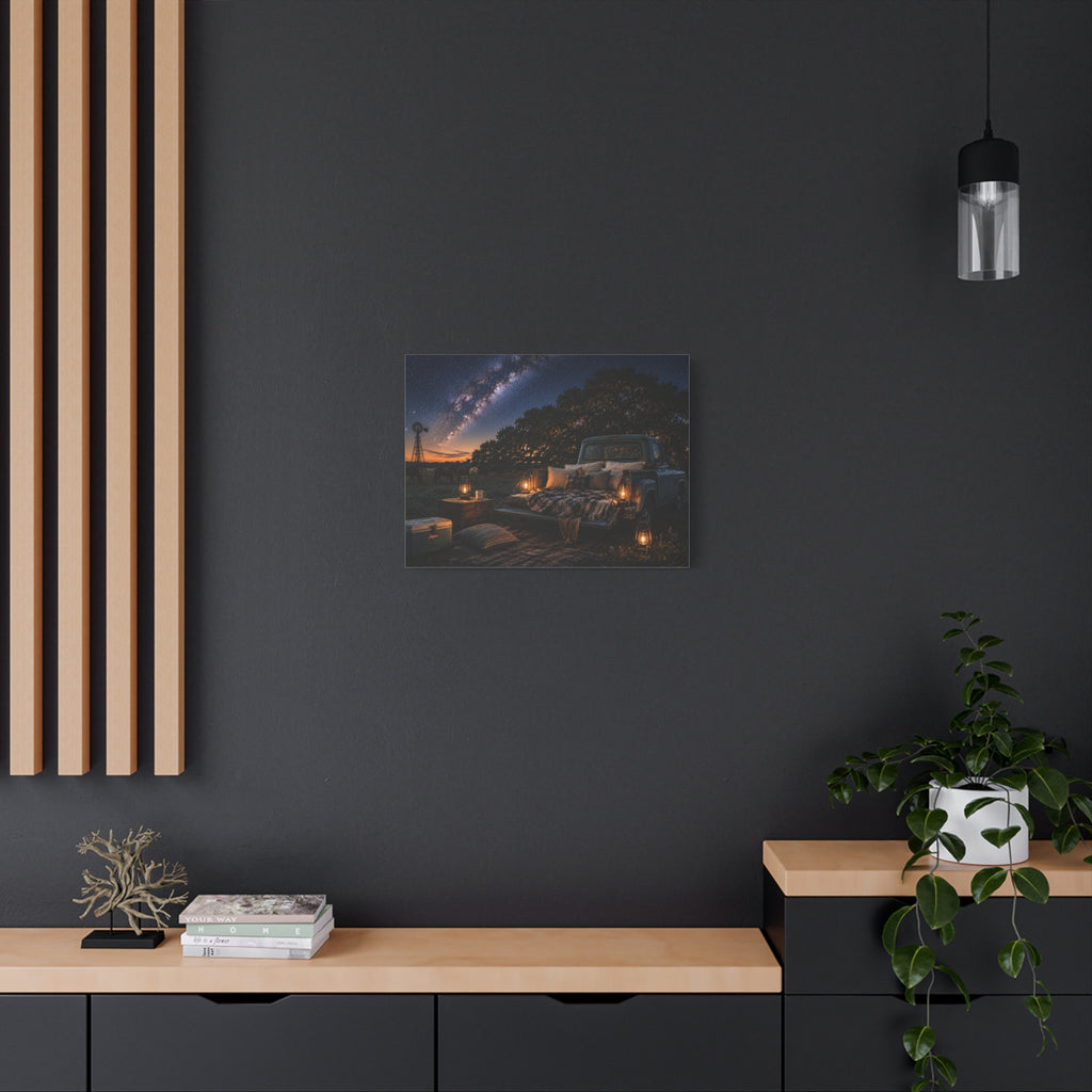 A square Starry Night canvas print showing a cozy outdoor bed beside a vintage pickup in a country field, with lanterns, pillows, blankets, windmills, and the Milky Way overhead, displayed on a dark charcoal wall above a light wood console with books and a plant.
