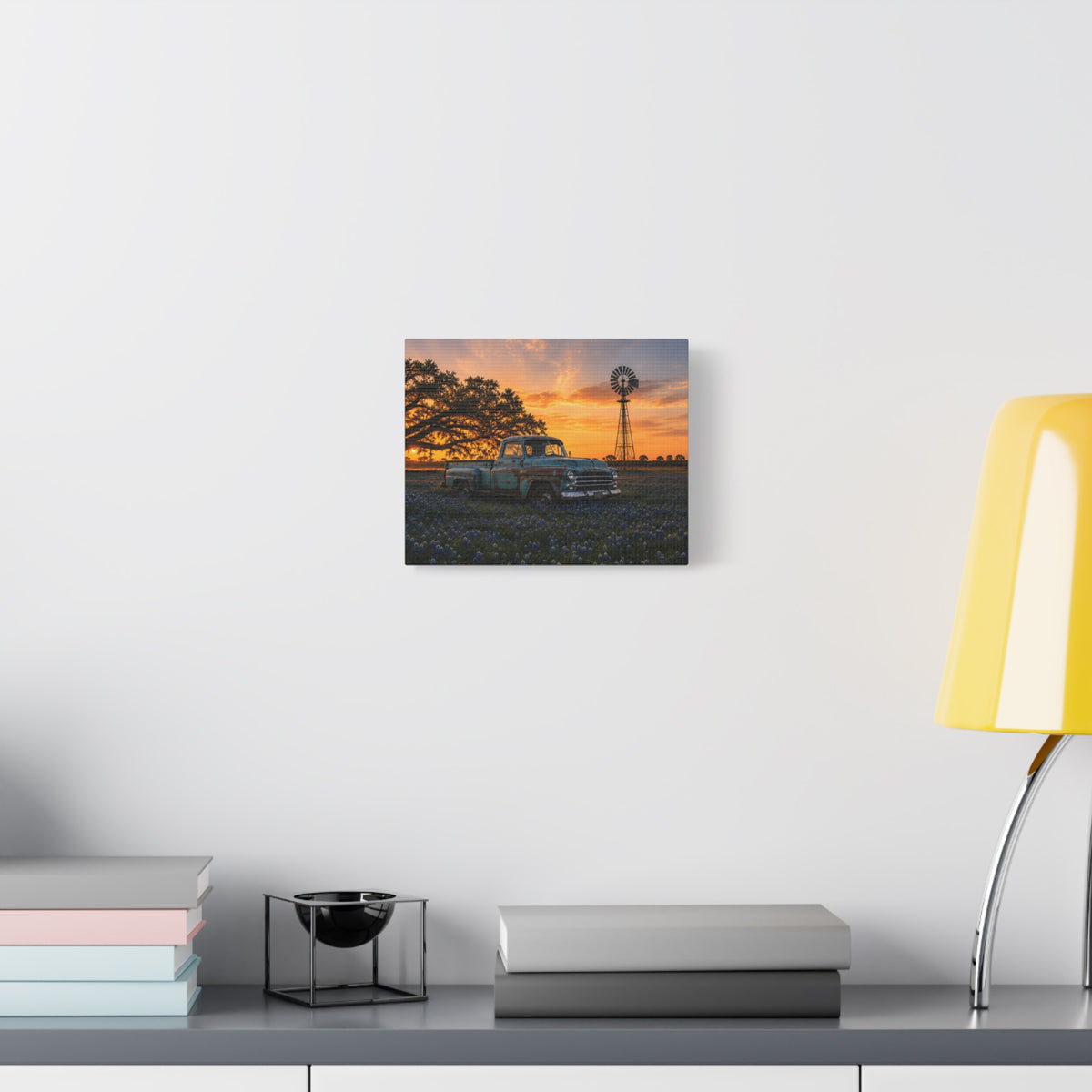 A rectangular The Windmill canvas print showing a weathered blue vintage pickup in a field of bluebonnets at sunset, with a windmill and large oak tree in the background, displayed on a dark wall above wooden shelves with a plant and decor.