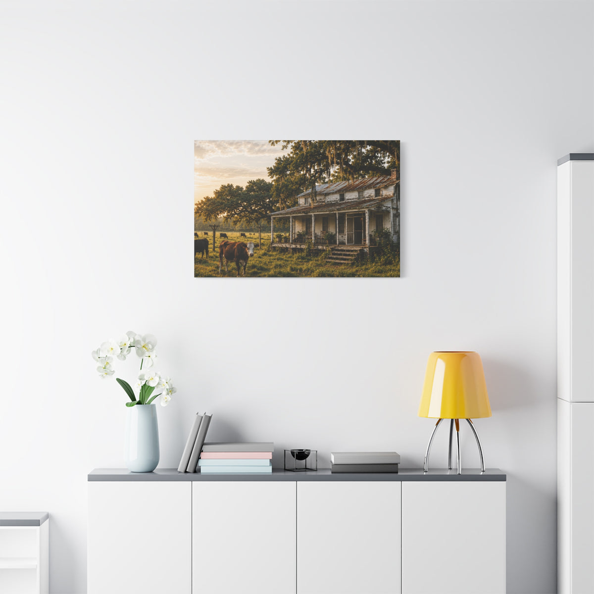 A The Good Old Homestead canvas print showing a weathered two-story farmhouse with a front porch in a grassy pasture, with a cow in the foreground, cattle, moss-draped oak trees, and warm sunset light, displayed on a white wall above a console with a yellow lamp, books, and white flowers.