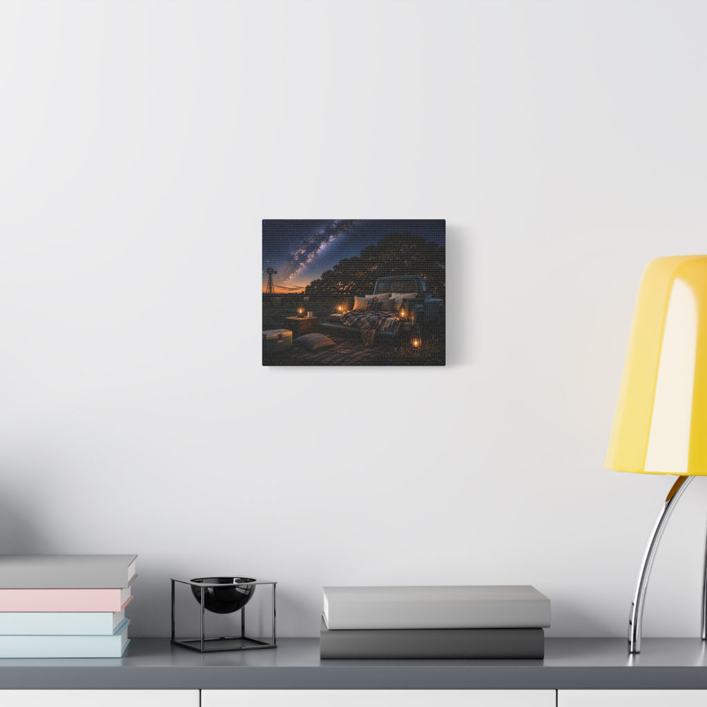 A square Starry Night canvas print showing a cozy outdoor bed beside a vintage pickup in a country field, with lanterns, pillows, blankets, windmills, and the Milky Way overhead, displayed on a white wall above books and a yellow lamp.
