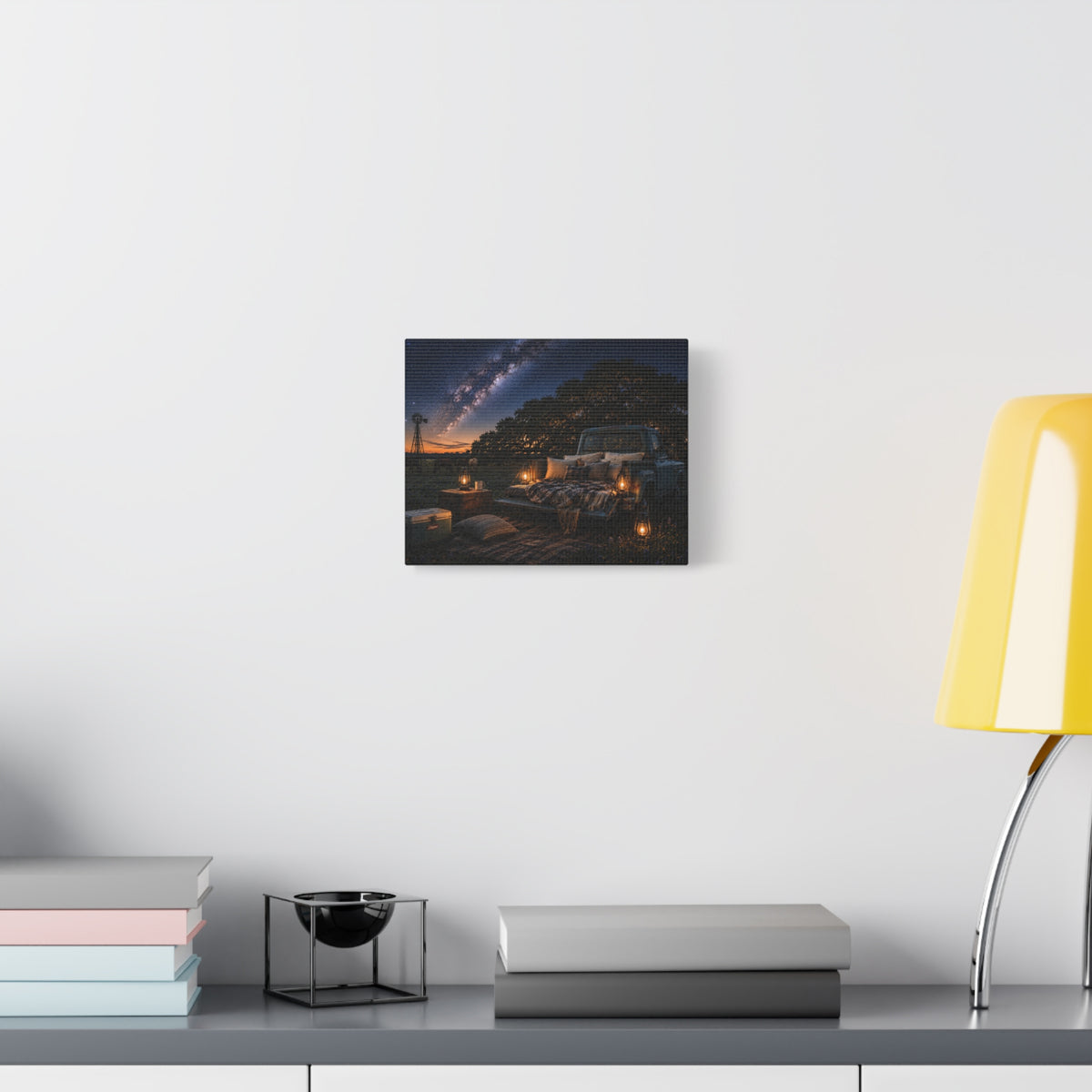 A square Starry Night canvas print showing a cozy outdoor bed beside a vintage pickup in a country field, with lanterns, pillows, blankets, windmills, and the Milky Way overhead, displayed on a white wall above books and a yellow lamp.