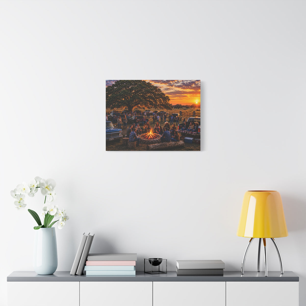 A square Campfire Stories canvas print showing friends gathered around a ranch campfire beneath a large oak at sunset, displayed on a dark gray wall above a wooden console with a plant and hanging light.